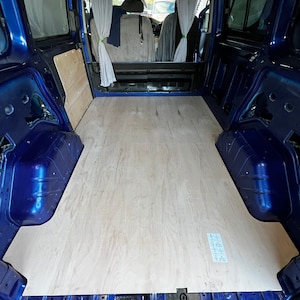 May include: Interior view of a blue van with a newly installed light wood floor. The van's side panels and rear door are visible, along with curtains and the front seats. The floor appears to be made of plywood.