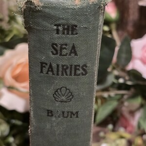 Op de afbeelding: Close-up van een vintage boekrug. De titel "THE SEA FAIRIES" is in zwart gedrukt op een verbleekte groene kaft. De naam van de auteur, "BAUM", staat onder een schelpenillustratie. De randen van het boek vertonen slijtage.