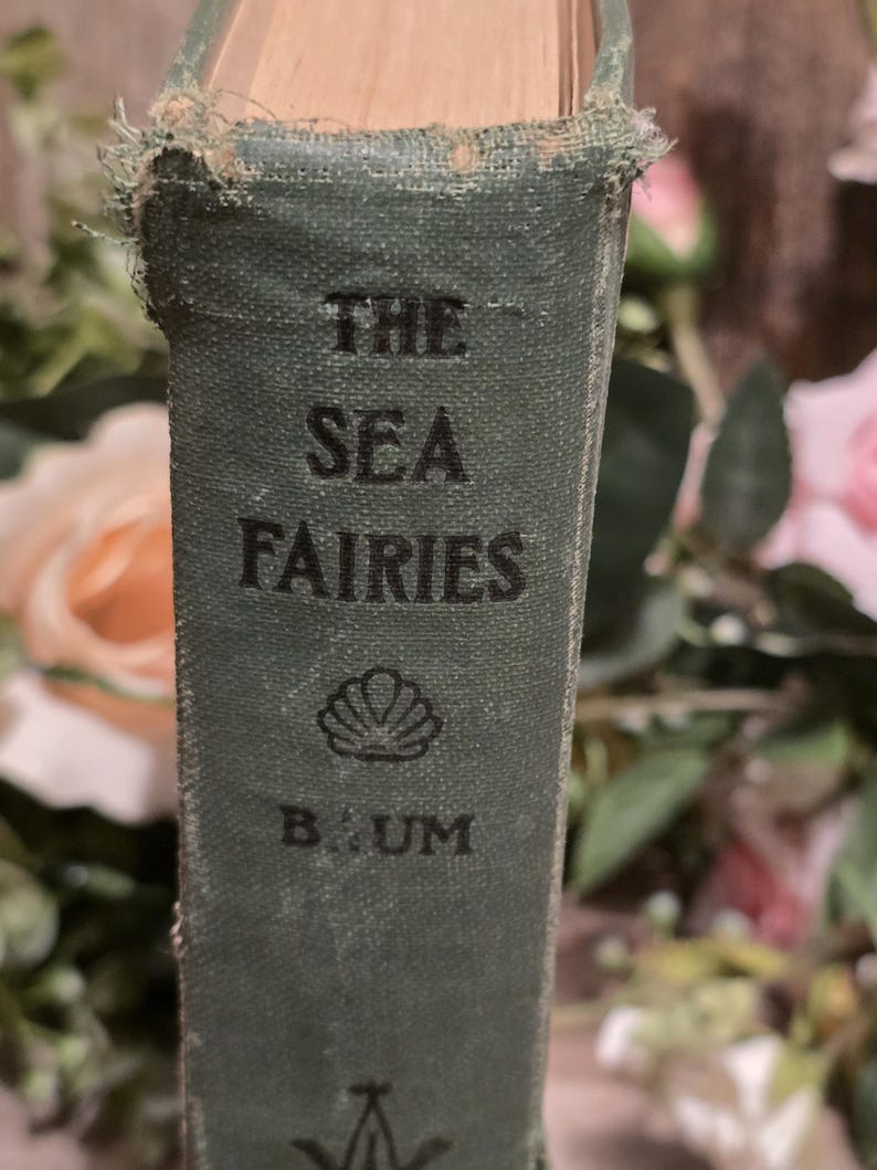 Op de afbeelding: Close-up van een vintage boekrug. De titel "THE SEA FAIRIES" is in zwart gedrukt op een verbleekte groene kaft. De naam van de auteur, "BAUM", staat onder een schelpenillustratie. De randen van het boek vertonen slijtage.