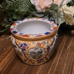 May include: A round, ceramic planter with a cream-colored base and intricate floral and bird designs. The rim is adorned with blue, orange, and pink floral patterns. The planter is likely for indoor use and adds a touch of elegance to any space.