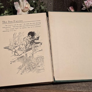 Op de afbeelding: Open vintage boek getiteld "The Sea Fairies". De linkerpagina bevat tekst en een illustratie van een man. De rechterpagina is leeg. Het boek ligt open op een houten oppervlak met bloemaccenten.