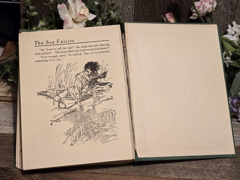 Op de afbeelding: Open vintage boek getiteld "The Sea Fairies". De linkerpagina bevat tekst en een illustratie van een man. De rechterpagina is leeg. Het boek ligt open op een houten oppervlak met bloemaccenten.