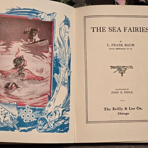 Op de afbeelding: Open boek met de titel "THE SEA FAIRIES" van L. Frank Baum. De linkerpagina bevat een kleurenillustratie van zeemeerminnen in het water, met een boot en een persoon. De illustratie is omlijst met blauwe en rode decoratieve elementen.