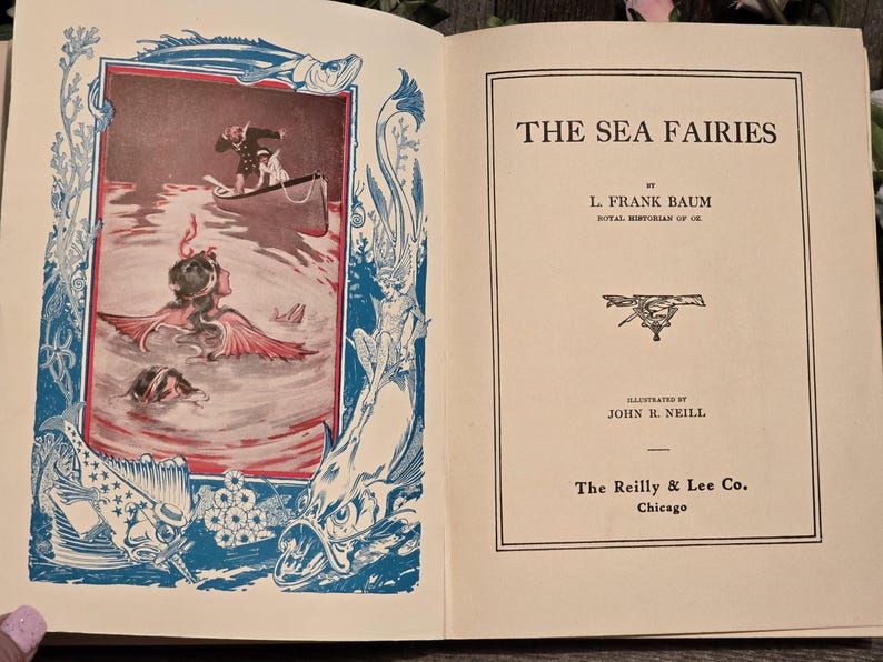 Op de afbeelding: Open boek met de titel "THE SEA FAIRIES" van L. Frank Baum. De linkerpagina bevat een kleurenillustratie van zeemeerminnen in het water, met een boot en een persoon. De illustratie is omlijst met blauwe en rode decoratieve elementen.