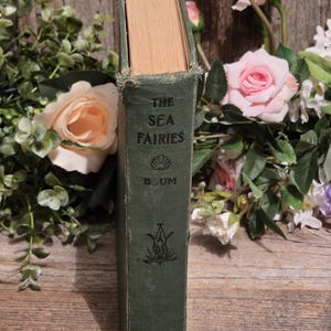 Op de afbeelding: Een vintage hardcover boek getiteld "The Sea Fairies" van L. Frank Baum. Het boek heeft een versleten groene kaft met gouden letters en een decoratief embleem. Omringd door roze en perzikrozen en groen.