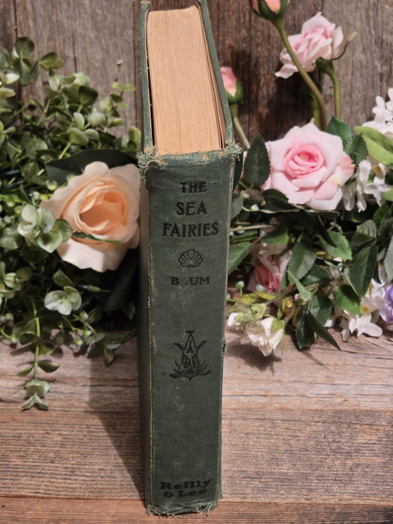 Op de afbeelding: Een vintage hardcover boek getiteld "The Sea Fairies" van L. Frank Baum. Het boek heeft een versleten groene kaft met gouden letters en een decoratief embleem. Omringd door roze en perzikrozen en groen.