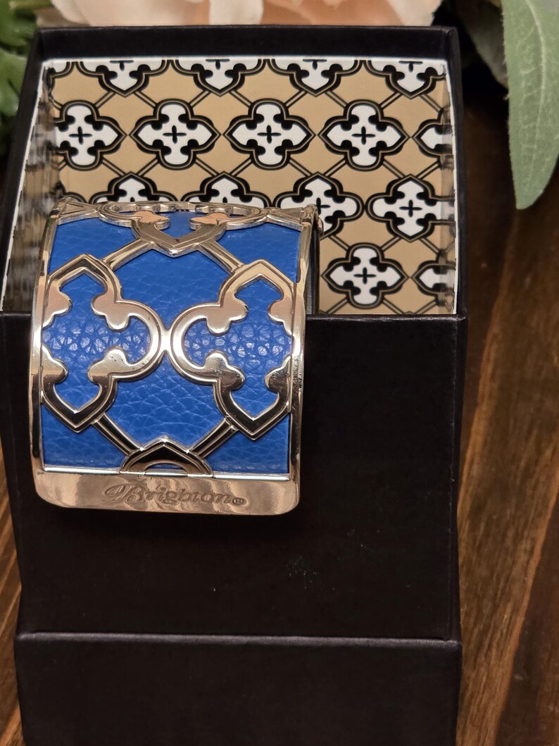 Pu&ograve; includere: Un bracciale a polsino blu e argento con un design geometrico, presentato in una scatola nera. Il bracciale presenta un centro blu strutturato e dettagli argentati. La scatola ha un interno a fantasia. La parola "Brighton" &egrave; incisa sul bracciale.