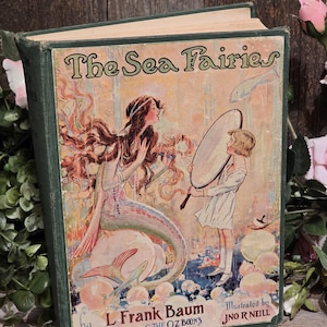 Op de afbeelding: Vintage hardcover boek getiteld "The Sea Fairies" door L. Frank Baum, ge&iuml;llustreerd door J.N.R. Neill. De omslag toont een zeemeermin en een kind met een spiegel. Het boek heeft een groene kaft en verouderde pagina's.