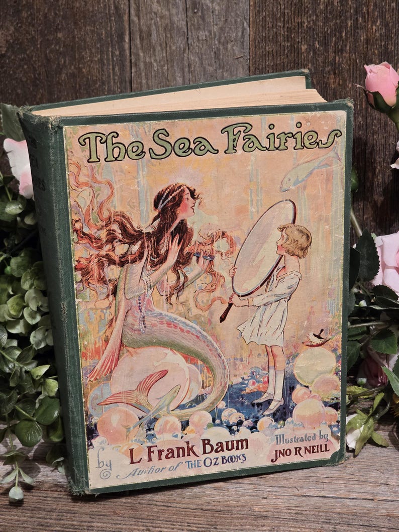 Op de afbeelding: Vintage hardcover boek getiteld "The Sea Fairies" door L. Frank Baum, ge&iuml;llustreerd door J.N.R. Neill. De omslag toont een zeemeermin en een kind met een spiegel. Het boek heeft een groene kaft en verouderde pagina's.
