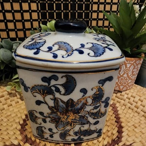 May include: A white ceramic jar with a lid, adorned with blue floral patterns and gold accents. The jar has a dark blue knob on top and is set against a natural woven background. The floral design includes stylized flowers and leaves.