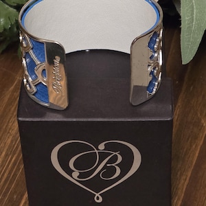Pu&ograve; includere: Un bracciale a polsino argentato con dettagli blu e bianchi. Il bracciale presenta un design traforato con sezioni blu e il nome del marchio "Brighton" inciso. &Egrave; esposto su una scatola nera con un logo a forma di cuore. Circonferenza interna di circa 17 cm.