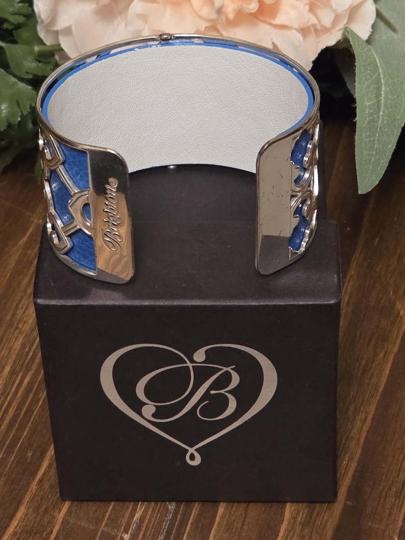Pu&ograve; includere: Un bracciale a polsino argentato con dettagli blu e bianchi. Il bracciale presenta un design traforato con sezioni blu e il nome del marchio "Brighton" inciso. &Egrave; esposto su una scatola nera con un logo a forma di cuore. Circonferenza interna di circa 17 cm.