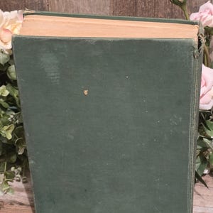 Op de afbeelding: Een vintage boek met een donkergroene kaft en vergeelde pagina's. De kaft van het boek vertoont tekenen van slijtage, met een enigszins versleten uiterlijk. Roze rozen en groen zijn zichtbaar op de achtergrond, wat wijst op een rustiek of antiek thema.