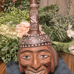 May include: A brown ceramic vessel with a smiling face design. The vessel has a cylindrical neck and a rounded body. The face is painted with detailed features, including eyes, a mouth, and decorative patterns. The vessel is likely a decorative item or a piece of art.