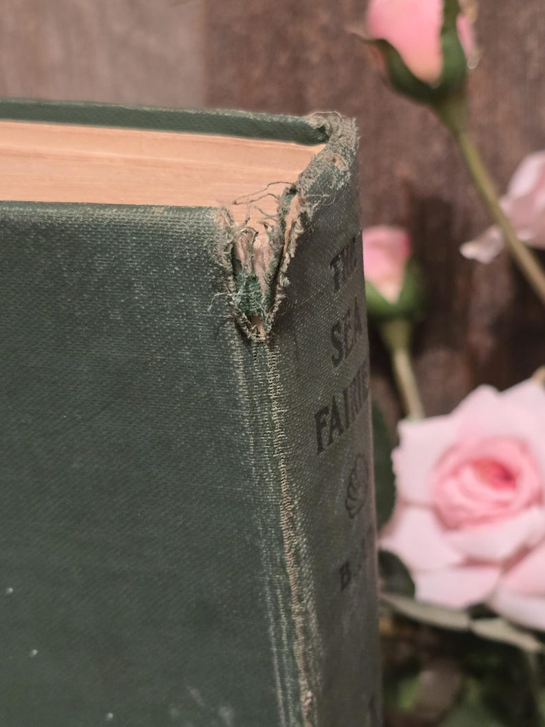 Op de afbeelding: Close-up van een vintage boek getiteld "The Sea Fairies" van L. Frank Baum. Het boek heeft een versleten, donkergroene kaft met zichtbare slijtage aan de hoeken. De pagina's zijn vergeeld en de titel is gedeeltelijk zichtbaar.