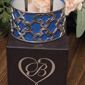 Pu&ograve; includere: Un bracciale a polsino in pelle blu con un design a filigrana argentata. Il bracciale ha un interno in pelle bianca ed &egrave; esposto su una scatola nera con un cuore e il logo della lettera "B".