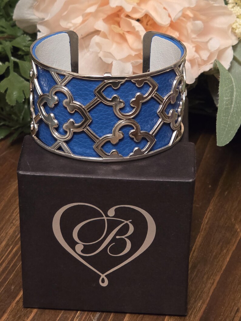 Pu&ograve; includere: Un bracciale a polsino in pelle blu con un design a filigrana argentata. Il bracciale ha un interno in pelle bianca ed &egrave; esposto su una scatola nera con un cuore e il logo della lettera "B".