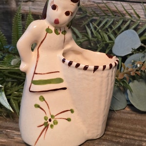 May include: A ceramic figurine of a girl holding a white planter with brown and white stripes. The girl is wearing a green and brown dress with a green floral design. The planter is decorated with a brown and white striped pattern.