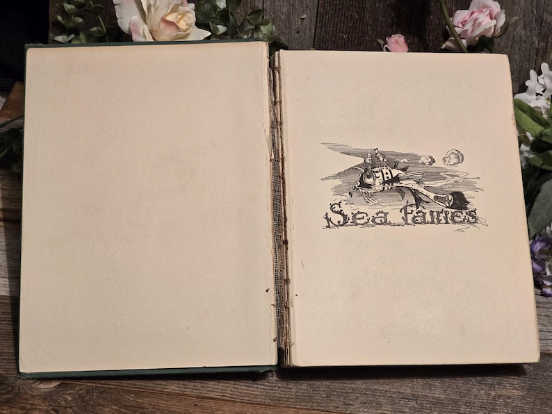Op de afbeelding: Een open vintage boek met verouderde, cr&egrave;mekleurige pagina's. De rechterpagina bevat een zwart-wit illustratie van een visachtig wezen met de tekst "Sea Fairies" eronder. De rug van het boek is zichtbaar en de kaft is donkergroen.