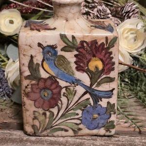 May include: A small, square ceramic vase with a crackled glaze. The vase is decorated with a blue bird, red and blue flowers, and green leaves on a white background.