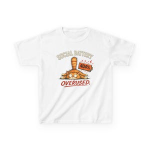 Puede incluir: Una camiseta blanca con un gráfico de un gato atigrado naranja tumbado boca abajo con la cola erguida. Junto al gato hay un icono de batería naranja con la etiqueta 404%. El texto dice SOCIAL BATTERY arriba y OVERUSED. en rojo abajo.