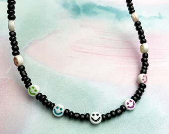 May include: A black beaded necklace with white smiley face beads. The smiley faces have different coloured outlines, including green, blue, and purple.