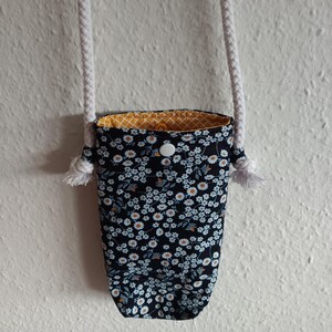 Könnte beinhalten: Eine kleine schwarze Stofftasche mit einem weißen Blumenmuster und gelbem Futter. Die Tasche hat einen weißen Knopfverschluss und einen weißen Kordelriemen.