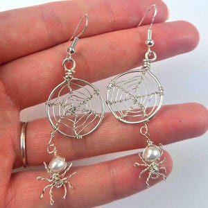 May include: Silver-toned wire earrings featuring a spiderweb design within a circular frame. Each earring has a dangling spider charm with a pearl body. The earrings have a hook closure.