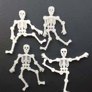 May include: Four white plastic skeleton figures posed in different positions on a black background. The skeletons are articulated at the joints and have a simple design.