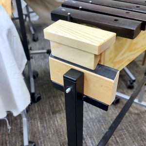 Puede incluir: Un xilófono de madera con teclas negras y un soporte de madera con un soporte de metal negro. El xilófono tiene una tapa de madera que se puede utilizar para guardar las baquetas.