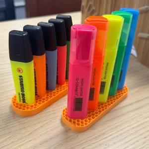 May include: A set of eight highlighter pens in a variety of colors, including yellow, orange, pink, red, green, blue, and purple. The pens are arranged in a honeycomb-shaped orange plastic holder. The text on the pens reads "STABILO BOSS ORIGINAL" and "HIGHLIGHTER Q-CONNECT+ STABILO WATERPROOF".