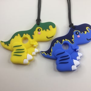 May include: Two dinosaur-shaped teething necklaces. One is yellow with green accents, the other is blue with navy accents. Both have a black cord and a hole in the centre. The dinosaurs have white feet and pink cheeks.