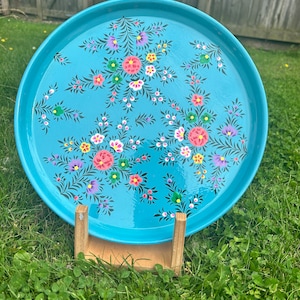 May include: A round, light blue metal tray featuring a hand-painted floral pattern with pink, purple, yellow, and green blossoms and dark green leafy sprigs. The tray is supported by a light-coloured wooden stand, displaying a smooth, glossy finish.