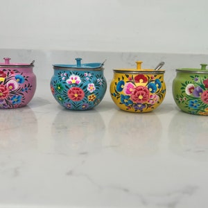 May include: Four small, round, hand-painted ceramic containers with lids and spoons. The containers are decorated with colourful floral patterns in pink, blue, yellow, and green.
