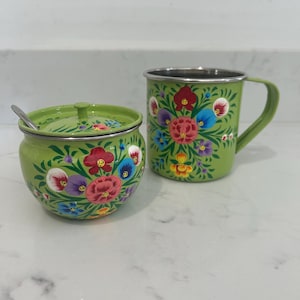 May include: A green enamel sugar bowl with a lid and a matching mug. Both are decorated with colourful floral patterns in red, pink, blue, and yellow. The mug has a silver rim and handle. The sugar bowl has a small spoon.