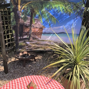 May include: A tropical beach scene printed on a fabric tapestry, featuring a hut on a white sandy beach with palm trees and turquoise water. The tapestry is hanging on a wooden frame in a garden setting.