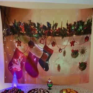 May include: A Christmas-themed tapestry featuring a fireplace mantel decorated with garland, lights, and ornaments. Three red stockings hang below, with a reindeer, stripes, and a geometric pattern. A small Christmas tree and other decorations are on the table.