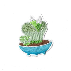 May include: An embroidered patch featuring a light green cactus with darker green spots and a small bright green sprout, sitting in a brown soil base within a rounded, bright blue ceramic-style pot with three small feet at the bottom.