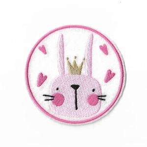 May include: A circular embroidered patch featuring a light pink rabbit face with a gold crown, black nose and whiskers, and pink cheeks. The design is set against a white background with four small pink hearts, all enclosed by a pink border.