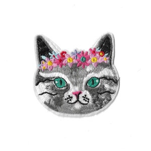 May include: An embroidered patch shaped like a cat head with gray and white fur, bright green eyes, and a pink nose. The cat is wearing a colorful floral crown featuring small pink, yellow, blue, and red flowers across the top of its head.
