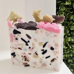 May include: A square bar of handmade soap with a white base, speckled with black, brown, and pink fragments. The top is decorated with small, colorful mushroom-shaped embellishments in shades of beige, pink, and brown. The soap has a unique, textured appearance.