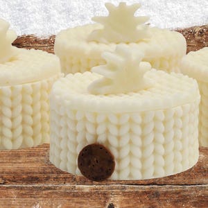 May include: Ivory-colored, round decorative boxes with a knitted pattern and a star-shaped accent on top. A small, round, brown accent is on the side of one box. The boxes are arranged on a wooden surface with a snowy background.