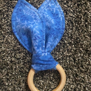 May include: A blue and white patterned fabric teether with a wooden ring. The teether is tied in a knot at the top of the ring.
