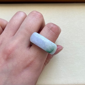 Peut inclure: Une bague rectangulaire en jade aux tons bleu clair et vert. La bague présente une surface lisse et polie, mettant en valeur les motifs naturels de la pierre. La bague est portée au doigt.