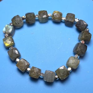 May include: A beaded bracelet featuring faceted, square-cut gemstones in shades of green, gray, and yellow. The stones are interspersed with small, silver-colored beads. The bracelet is displayed on a blue surface.