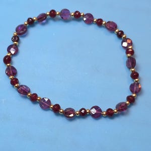 May include: A beaded bracelet featuring alternating faceted purple amethyst beads and smaller deep red garnet beads, interspersed with tiny polished gold-tone metal spacer beads arranged in a continuous circular pattern on a flexible cord.