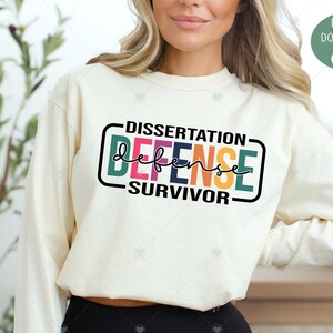 Könnte beinhalten: Ein weißes Sweatshirt mit dem Text "DISSERTATION DEFENSE -SURVIVOR- " in einem Regenbogen-Farbschema. Der Text ist in einer fetten, klobigen Schrift.