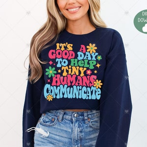 Puede incluir: Una sudadera azul marino con un gráfico colorido que dice "It's a good day to help tiny humans communicate". El gráfico presenta flores y estrellas.