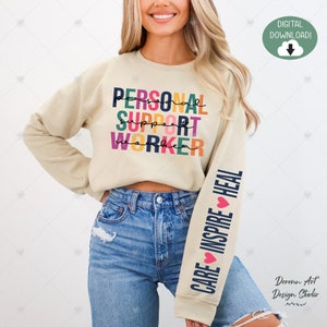Peut inclure: Un sweat-shirt beige avec le texte "PERSONAL SUPPORT WORKER" en couleurs arc-en-ciel sur le devant et le texte "CARE INSPIRE HEAL" avec des cœurs sur la manche.