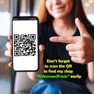 May include: A person holding a smartphone displaying a QR code. The text on the screen reads "Don't forget to scan the QR to find my shop "RaberoanPrints" easily."